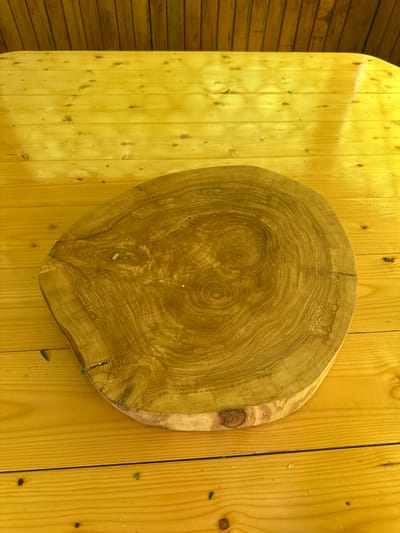 A wooden table with a distinctive circular wood slice or cutting board placed on it. The table is made of light-colored wood planks. The wood slice has prominent growth rings and a natural, irregular edge. The background consists of vertical wooden slats, likely part of a wall or fence. The scene is illuminated, suggesting an outdoor or well-lit indoor setting. There are no people or animals present. The location is Yazykova Pyatina, Russia.