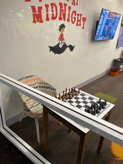 The scene shows a chessboard set up on a wooden table in a room in Indooroopilly, Australia (latitude: -27.501, longitude: 152.972). The table is situated on a carpeted floor with a patterned design. A colorful patterned chair sits to the left of the table, facing the board. Behind the table and chair, a white wall with a black and white cartoon illustration of a boy riding a wolf is seen. The inscription "at Midnight" is written in red across the top of the wall. A flat-screen television with a red "O-Post" logo is mounted on the wall, showing a news program. A small, brown, plastic storage bin can be seen to the right of the table, against the wall. The room is lit by overhead lighting, casting soft shadows. The overall environment is a modern interior space.