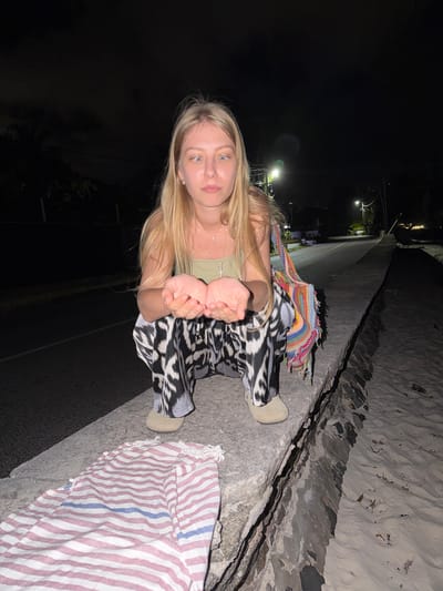 In Mare Anglaise, Seychelles, at night, a young woman with blonde hair is crouched on a concrete barrier next to a road. She is wearing a light green tank top, black and white patterned pants, and tan slip-on shoes. A colorful, fringed crocheted bag is draped over her right shoulder. Her hands are cupped together in front of her. A pink and white striped fabric is laid out on the barrier in the foreground. The background shows a dimly lit street with streetlights, palm trees, and other vegetation, suggesting a tropical location. The sky is dark with some clouds.