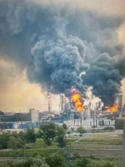 The image depicts a large industrial facility engulfed in flames and thick black smoke. The fire appears to be a significant event, with bright orange flames visible amidst the smoke. The industrial complex has numerous structures, including what appear to be towers and pipelines, typical of a chemical or oil refinery. In the foreground, there is greenery, suggesting the facility is located in or near an area with vegetation, possibly a park or agricultural land. The sky is obscured by the dark plume of smoke, though a lighter, possibly orange or yellow hue is visible near the horizon, suggesting it might be dusk or dawn. There is no visible indication of people or specific damage to surrounding areas beyond the immediate vicinity of the fire. The location is identified as the city of Sof’ino, Russia.