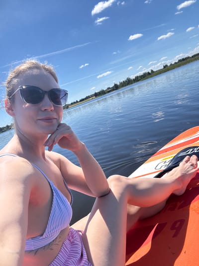 In Lendži, Latvia, a woman is captured in a self-portrait while paddleboarding on a body of water under a bright, partly cloudy sky. She is positioned on the orange paddleboard, with her legs extended. She is wearing a striped bikini and sunglasses. The paddleboard has visible markings. The water is calm, reflecting the blue sky and distant treeline, suggesting a serene outdoor environment.