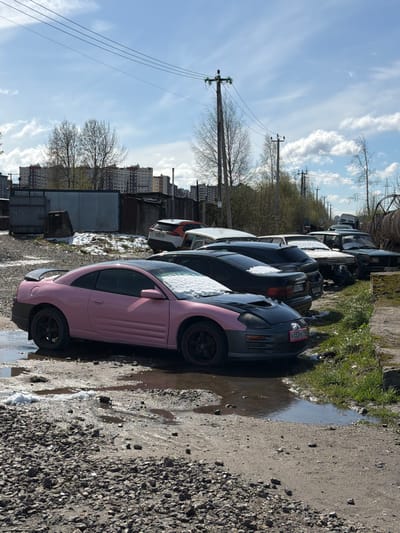 The image captures an outdoor scene in Lavriki, Russia, depicting a row of parked cars on a gravel and dirt lot. The perspective is from slightly below, looking along the line of vehicles. In the immediate foreground, a striking pink and black two-door coupe, possibly a Mitsubishi Eclipse, is prominently displayed, its hood and roof partially covered in patches of snow. A Russian license plate, "У 433РЕ 198," is visible on its front bumper.
Behind the pink car, several other older-model cars, mostly dark-colored sedans and coupes, are parked in succession, also bearing traces of snow on their surfaces. The ground beneath and around the cars is a rough mix of gravel, dirt, and mud, interspersed with numerous puddles of water that reflect the bright sky. A strip of green grass and sparse, bare-branched trees runs along the edge of the lot, separating it from the line of utility poles and overhead power lines that stretch across the mid-ground.
In the distance, multi-story apartment buildings are visible under a bright, partly cloudy sky, featuring broad streaks of white clouds against a vibrant blue. The presence of both snow and significant puddles suggests that the image was taken during early spring, likely during a period of thaw. The time of day is clearly daylight, with shadows indicating a sunlit scene. No people are present, and there is no specific activity taking place; the cars appear stationary, lending a quiet and somewhat desolate atmosphere to the scene.