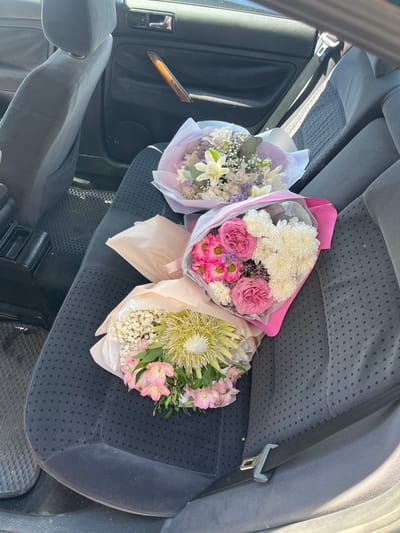Three flower bouquets are arranged on the back seat of a car. The car interior appears to be dark gray fabric. The seat belt is buckled and visible. The image does not provide any information about the city of Kobryn, Belarus.