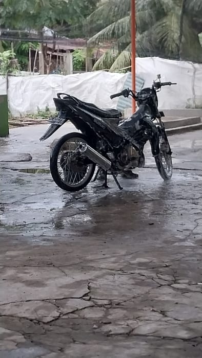 A black Suzuki underbone motorcycle is parked on its kickstand on a wet, cracked concrete surface in Mali Guyui, Indonesia. The motorcycle, oriented with its rear towards the viewer, is visibly saturated with water. A silver-colored exhaust pipe is prominent on the right side of the motorcycle, and its rear wheel features chrome spokes. The concrete ground is extensively cracked and covered with numerous puddles, reflecting light. In the background, white tarpaulins cover structures, with dense green foliage and palm trees visible above and behind them. A vertical orange pole stands in the mid-ground, to the right of the motorcycle's handlebars. No individuals are present in the scene.