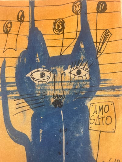 The image displays a close-up view of a fabric with a printed design. The design features a blue abstract cat-like figure on a tan background with vertical lines. There are also black ink drawings of musical notes and a box containing the text "AMO GATO". The style is whimsical and childlike. The fabric appears to be a light-weight material, possibly cotton or linen, with a visible woven texture. The tan background suggests a warm, possibly outdoor setting, and the "AMO GATO" text, which translates to "I love cat" in Spanish, indicates a playful theme. Given the location context of Calp, Spain, it is plausible this is a decorative item like a tea towel or a piece of fabric art related to local culture or art. The colors are vibrant, with the blue of the cat standing out against the warm tan. There are no people or specific environmental details like time of day or weather visible, focusing solely on the printed design.