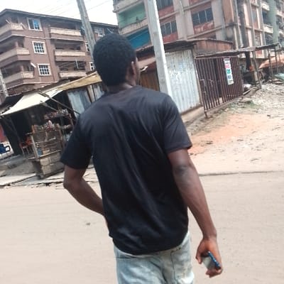 The image depicts an urban street scene in Awada, Nigeria. A male subject, viewed from behind, occupies the foreground, slightly off-center to the right. He has short, dark, curly hair and dark skin, wearing a black short-sleeved t-shirt and light-colored, possibly faded, jeans. His left hand is positioned towards his hip, while his right hand holds a dark-colored smartphone. The ground beneath him is an unpaved or dirt road of reddish-brown earth.

In the background, on the left, a multi-story brown residential building with multiple balconies and white-framed windows is visible. Adjacent to the road, several small, rudimentary commercial structures made of corrugated metal are present. One on the immediate left appears to be an open stall displaying various goods. Further to the right, another multi-story building with green and faded reddish-brown sections is visible. A prominent utility pole stands in the mid-ground. Near the pole, another corrugated metal structure features a closed metal gate adorned with several posters or flyers. The sky is a bright, uniform light tone, suggesting an overcast or hazy day. No direct interactions between individuals are discernible.
