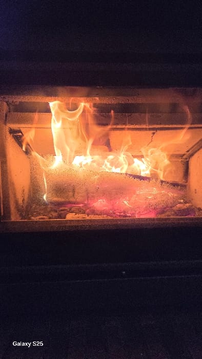 The scene depicts an active wood fire contained within a glass-fronted stove or fireplace insert in Ottonville, France. Several logs of wood are actively burning, producing bright orange and yellow flames that rise from glowing red embers. The fire is viewed through a glass panel, which exhibits a fine speckling of particulate matter or condensation. The immediate environment outside the firebox is dark, indicating a low-light indoor setting. No individuals are present in the image. The text "Galaxy S25" is visible in the lower-left corner.