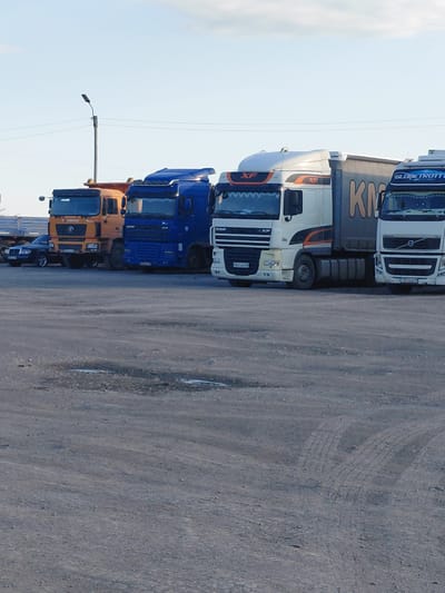 In the city of Molodezhny, Kazakhstan, a row of parked semi-trailer trucks and a dark-colored passenger car are visible on a paved lot under a clear sky with a few clouds. The trucks are of various colors, including orange, blue, white, and white with a gray trailer. The truck on the far left is orange and appears to be a dump truck. Next to it is a blue semi-trailer truck. To the right of that is a white semi-trailer truck with a gray trailer, and further to the right is another white semi-trailer truck. The passenger car, a dark sedan, is parked in front of the orange truck. A lamppost with wires is visible in the background. The ground in the foreground is a rough, gray surface with some patches of wetness and tire tracks. The lighting suggests it is daytime, possibly late afternoon or early evening.