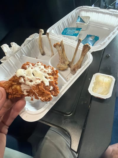 A person's hand is holding a fried chicken wing that has been drizzled with a white sauce. The wing is resting in a white plastic takeout container filled with chicken bones. Another identical container is to the right, containing small packets of condiments. The scene appears to be indoors, possibly in a car or a dimly lit room, with a dark background and a dark surface in the foreground.