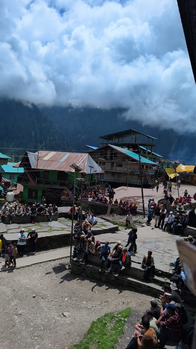 In Malana, India, a bustling gathering of approximately 100 people is captured on a paved, uneven courtyard. The scene is dominated by traditional Himachali architecture, with several multi-story wooden buildings featuring distinct corrugated metal roofs in various states of repair. One prominent green building displays a rusty, colorful roof and a set of stairs leading to an upper level. Adjacent to it, another building has a turquoise roof and balconies. Further in the background, a taller structure with multiple levels and a similar turquoise roof is visible, along with makeshift yellow awnings. The sky is dramatic, with large, soft white clouds obscuring a dark, forested mountain range in the distance.
The people are primarily seated on stone walls and steps that border the courtyard, while others stand or walk through the open spaces. Many are dressed in traditional Himachali attire, including distinctive caps. There's a sense of communal activity, with individuals engaged in conversations and observing their surroundings. Some are holding mobile phones, suggesting the presence of modern technology within this traditional setting. The ground is a mixture of stone paving, packed dirt, and patches of green grass. The overall impression is of a vibrant, communal gathering in a picturesque mountain village.