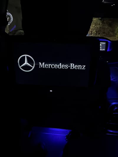 The image depicts the interior of a Mercedes-Benz vehicle at night in the city of Dragor, Bulgaria. The central focus is a rectangular screen displaying the Mercedes-Benz logo, a silver three-pointed star within a circle, to the left of the white text "Mercedes-Benz." A minute, barely legible symbol or text, possibly "ON," is visible below the brand name on the screen. Below this main screen, a horizontal strip of bright blue ambient light illuminates the lower section of the dashboard or console.
To the upper left, a dark fabric object, possibly a seatback or cover, is partially visible, featuring a circular white graphic resembling a speedometer or gauge with white markings and a faint blue accent.
To the upper right, a blurred view through a window shows indistinct, textured shapes, possibly foliage or a wet surface outside. Below this, a smaller illuminated display module is visible, encased in a prominent blue light. This module contains a small screen with faint white text or symbols, resembling a "D" or "P," likely indicating a gear selection or status. The overall interior is dark, with prominent blue ambient lighting accentuating various elements within the cabin, including the floor area visible at the bottom of the frame.
