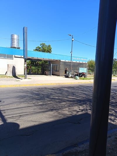 The image depicts an exterior view of the "Vice Alte. Montes" train station in Don Torcuato, Argentina, under a clear blue sky. The station building, primarily white or light gray with a prominent light blue fascia, features a metal water tank on its roof and a tall, slender vertical sign column. The entrance is marked by black metal gates.

On the left side of the sidewalk, a large, weathered tree stump is visible. Further right, a red bicycle is leaning against the station's entrance fence. A single individual, dressed in dark attire, stands on the sidewalk near a gray trash bin or collection cart, adjacent to the building's right section, which features horizontal slatted panels. To their right, a dark-colored motorcycle is parked on the sidewalk, with a green bag resting on the ground nearby. A small green informational sign with text and graphics is partially visible next to the motorcycle.

The scene includes an asphalt street with visible cracks and yellow-painted curbs. Long shadows, indicative of strong sunlight, are cast across the street and sidewalk, originating from unseen trees and utility poles. A black utility pole, equipped with a streetlight and a security camera, is positioned in the mid-ground. The right side of the frame is partially obscured by a dark gray, vertical pole. Green trees and foliage are present in the background, behind the station structure and a fence.