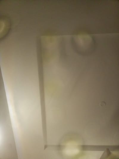 The image shows a ceiling with a recessed area and some subtle lighting effects or reflections. The ceiling appears to be a uniform off-white or light beige color. There is a distinct rectangular recess in the ceiling, with a visible edge. The rest of the ceiling is flat. There are some circular or oval shapes that appear to be light reflections or possibly water stains on the surface of the ceiling, particularly in the recessed area and towards the top left of the image. One small circular fixture, likely a light or smoke detector, is visible on the right side of the flat ceiling. The overall lighting in the scene is dim and diffused. The image is taken from a low angle, looking upwards towards the ceiling. The location is Unknown, Unknown.