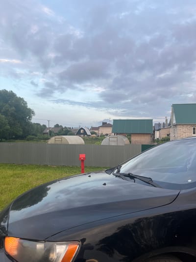 This media file captures a rural scene in Nikolskoye, Russia, likely during the late afternoon or early evening given the soft, diffused light and cloudy sky. The image is taken from the perspective of a person standing near a black car, which occupies the right side of the frame. The car's hood and headlight are visible, reflecting the cloudy sky and surrounding environment.
The scene is characterized by a collection of buildings with green corrugated roofs, typical of dachas or rural homes. Several of these structures are visible in the background, interspersed with trees and vegetation. A greenhouse with a transparent roof is also present. A red mailbox stands prominently on the left side of the frame, near a green metal fence that separates the yard from the buildings and further greenery.
The weather appears overcast with dramatic, textured clouds filling the sky. The ground is covered in green grass, suggesting a temperate climate and possibly summer or late spring. There is no visible text in the image. The overall mood is peaceful and tranquil, evoking a sense of rural solitude. The composition, with the car in the foreground and the village in the background, provides a glimpse into the daily life or landscape of this Russian locality.