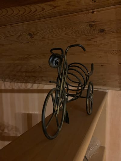 A metal, bicycle-shaped wine rack is positioned on a wooden shelf. The wine rack is black and has three wheels, a basket-like structure in the middle to hold bottles, and a handlebars. The background consists of wooden planks. The scene appears to be indoors, likely in Mepiskalaki, Georgia, though the specific location within the city is not determinable from the image.