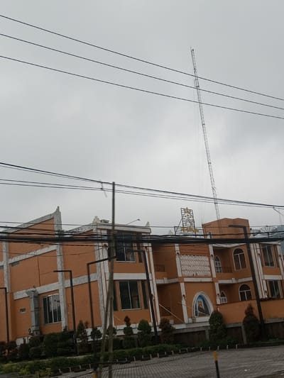 A multi-story building with orange facade and white trim sits beneath a cloudy sky. A tall communications tower rises behind it. Overhead power lines crisscross the view. Landscaping surrounds the base of the building, with small trees and bushes arranged neatly. A religious figure is depicted in an alcove on the building's facade, and a sign with a crown adorns the top. The building's design features arched windows and decorative architectural elements.