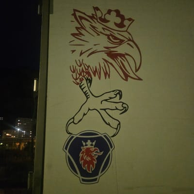 The image shows a tall, light-colored building with a dark silhouette of a stylized griffin's head and a claw on its upper portion. The griffin is depicted in dark lines with red accents on the crown and head. Below the claw is the Scania truck logo, a blue and white hexagonal emblem with a red griffin head inside. The building is adjacent to a dark, outdoor environment, possibly at night, with lights from other buildings visible in the background, suggesting an urban setting. The location is specified as Ventimiglia, Italy.