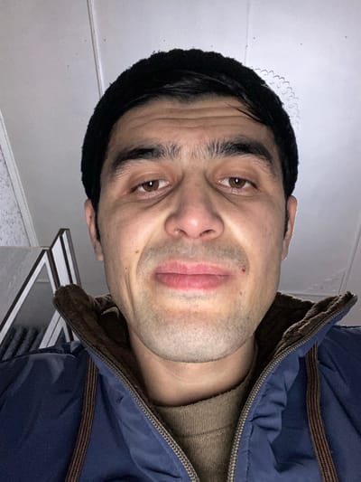 A man in a dark beanie and blue jacket is facing the camera. His brown eyes look directly at the viewer. He has dark eyebrows and a slight stubble. A small, reddish mark is visible near his lip. The background includes a white ceiling with decorative trim and a tiled surface with geometric black and white patterns.