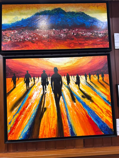 The image shows two framed abstract paintings displayed on a wooden wall. The top painting depicts a vibrant, textured landscape with a dominant blue and green mountain range against a yellow sky, set against a fiery red and orange foreground with white and dark speckles. The bottom painting is an impressionistic scene featuring silhouetted figures walking across a brightly colored, striped ground of orange, yellow, blue, and red. The figures cast long, dark shadows, suggesting a strong light source, likely a sunset or sunrise. A small, partially visible label on the right side of the top painting appears to be in Spanish and mentions "TORREJÓN DE ARDOZ, ESPAÑA," indicating the location of this display. The paintings are framed in black and are mounted on a wooden surface with vertical planks.