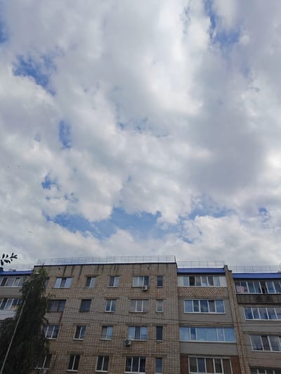 The image shows a view looking up at a multi-story apartment building in Kumertau, Russia. The building is constructed with light brown brick and features numerous windows, some of which are dark and reflect the sky. The top of the building is topped with a blue metal roof and a dark metal railing. A tall, leafy tree stands to the left of the building, partially obscuring some of its windows. The sky above is filled with a mix of white and gray clouds, with patches of blue sky visible. Several small, dark birds are scattered across the sky. The overall scene depicts a typical urban landscape on a cloudy day.