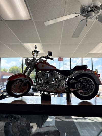 A red and silver model motorcycle is displayed on a shelf, with a ceiling fan and fluorescent lights visible above. The motorcycle is positioned in front of a large window that overlooks a street with trees and buildings. An "EXIT" sign is visible through the window. The scene is indoors, likely in a store or showroom. The image is taken from a low angle, focusing on the motorcycle.