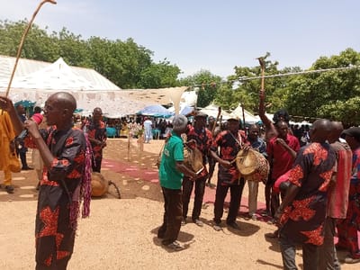 A group of men are playing drums and singing in an open-air space in Zum, Nigeria. The men are wearing brightly colored clothing. The drums are made of wood and have a round shape. The men are playing the drums with their hands and sticks. There is a microphone in front of one of the men, who is holding a long stick. There are other men standing in the background, watching the performance. The ground is covered in sand. There is a white tent and trees in the background. The scene is likely a celebratory event.
