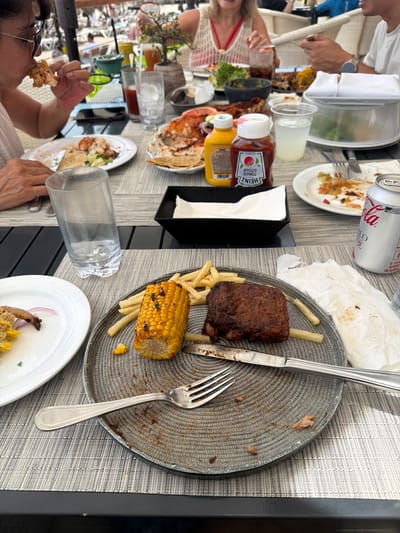 The image depicts a table setting in what appears to be an outdoor dining area. The scene includes remnants of a meal, suggesting that people have recently been eating at the table. The table is covered with a textured, possibly woven, placemat.

Present are multiple plates, some empty or containing food scraps. One plate has leftover ribs, french fries, and corn on the cob. There are also visible condiments, including ketchup and mustard bottles. Used napkins and empty beverage containers (e.g., a Coca-Cola can) are also present on the table.

Several people are visible, with some captured in the act of eating. One person is eating a piece of food with their hand. Other people are seated at the table, some partially obscured.

The setting appears to be outdoors. The location in the city of Las Jarretaderas, Mexico, is not directly depicted in the photo, therefore, this detail cannot be confirmed from this image.