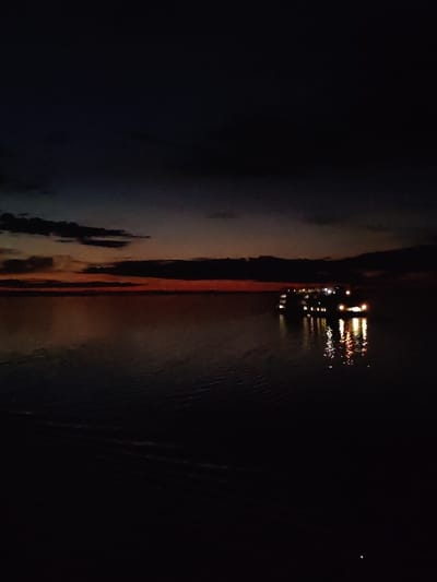 The image captures a nighttime scene at the former location of Mologa, Russia, now submerged by the Rybinsk Reservoir. A large vessel, likely a passenger or tourist boat, is illuminated with multiple lights, reflecting in the dark water. The sky is dark, with a gradient of orange and red at the horizon, indicating either a sunset or sunrise. A few dark clouds are present near the horizon. The overall environment suggests a still, open body of water.