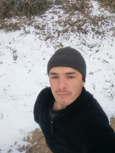 A man wearing a black winter coat and a grey beanie stands in a snowy landscape. Patches of snow cover the ground, with some bare patches visible. Behind him, a snow-covered slope or embankment rises, with bits of dry vegetation peeking through the snow. The man faces the camera with a neutral expression, a light mustache visible on his face. The overall scene suggests a cold, wintry day in a natural setting.