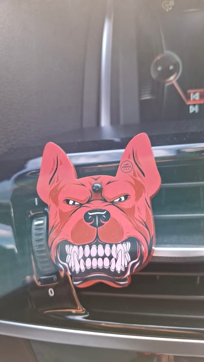 A red car air freshener shaped like a snarling dog's head is attached to the horizontal slats of a car's air conditioning vent. The air freshener has sharp teeth, angry eyes, and a small black stud on its forehead. The car's dashboard is visible in the background, with a portion of a digital display showing a circular gauge and playback controls. The scene is indoors, likely within a vehicle.