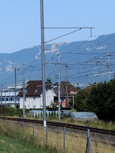 The image depicts a view of a residential area and surrounding landscape in Zuchwil, Switzerland, with a focus on infrastructure related to a railway. In the foreground, there is a railway track with a wire fence running along its edge. Beyond the fence, there is tall, dry grass.

In the middle ground, several buildings are visible. To the left, there is a modern, multi-story building with a blue and white facade. Next to it, a traditional Swiss house with a dark brown roof, white walls, and several windows is situated. To the right of this house, another building with a darker, pitched roof is partially visible, as are dense trees forming a backdrop.

The background is dominated by a large mountain range, with a clear, light blue sky above. Numerous overhead power lines and supporting poles, characteristic of a railway electrification system, crisscross the scene, creating a complex network of wires against the backdrop of the mountains. The overall impression is one of a semi-urban environment integrated with natural scenery and industrial infrastructure. There are no people visible in the image.