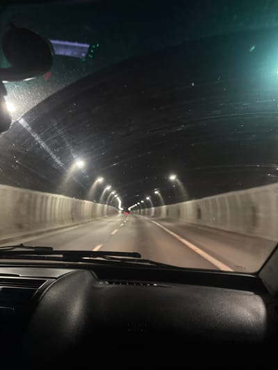 The image is a point-of-view shot from inside a vehicle traveling through a tunnel. The perspective is from the driver's seat, looking forward through the windshield. The tunnel interior is dark, with rows of lights lining the ceiling and walls, creating streaks of light and shadows. The road ahead is visible, with white lane markings. The dashboard of the car is in the foreground, obscuring the lower portion of the view. The scene appears to be a standard highway tunnel. There is no specific indication of the location being Dolna Vasilitsa, Bulgaria, within the image itself.