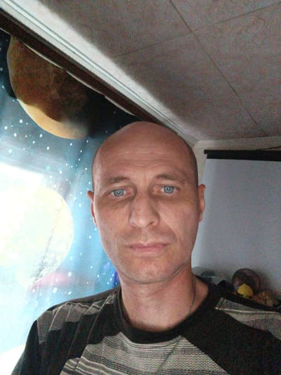 The image is a selfie taken by a bald, middle-aged Caucasian male with blue eyes. He is wearing a striped t-shirt and a silver chain necklace. The background appears to be a wall with a mural of outer space, featuring planets and stars. The lighting is dim, and the image quality is grainy. There is no information to indicate the location within Mokshan, Russia.