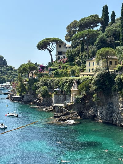 The image captures a sun-drenched, coastal scene in Portofino, Italy, showcasing the vibrant turquoise waters of the Ligurian Sea lapping against rocky cliffs. Lush green vegetation cascades down the hillsides, interspersed with elegant villas and homes.
In the foreground, the crystal-clear water reveals the seabed in shallow areas, while deeper sections are a rich, deep blue. Several small boats, including a white motorboat with an awning, are moored in the bay. Two figures are visible on the deck of one of the boats. Further out in the water, at least six individuals are swimming or floating, enjoying the refreshing sea.
The coastline is characterized by dramatic rocky formations, with terraced gardens and stone structures built into the cliffside. One notable structure is a small, ornate tower with a conical roof, situated on a rocky outcrop. Above the immediate coastline, several buildings are visible, including a prominent yellow villa with striped awnings and a larger white villa with arched windows, both nestled amongst the trees. The architecture reflects the distinctive style of the Italian Riviera.
The sky is a clear, bright blue, indicative of fair weather. The overall impression is one of idyllic Mediterranean beauty, a picturesque blend of natural landscape and human habitation.