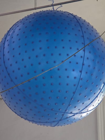 The media file shows a close-up, slightly angled view of a large, blue, spiked exercise ball. The ball is suspended by thin ropes, suggesting it might be part of an exhibit or a display. Two blue ropes are visible wrapped around the ball in a crisscross pattern, and a single, thin, tan-colored string or wire also diagonally crosses the ball's surface. The textured surface of the ball is covered in small, evenly spaced raised bumps. The background is a plain, light-colored wall with a white ceiling line visible at the top. There are no people or other prominent objects in the frame, and no discernible time of day or weather cues. The image appears to be taken indoors, given the lighting and the presence of a ceiling. The location context provided is Syrdarya, Uzbekistan, but there are no visual elements in the image that specifically indicate this location. There is no visible text in the media file.