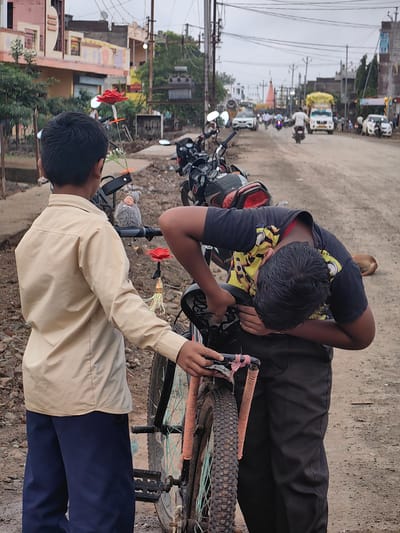 Two boys are standing next to a bicycle on a dirt road in the city of Padli, India. The boy on the left is wearing a tan collared shirt and dark blue pants. He is holding a stick with a red artificial flower and a small white flower. The boy on the right is wearing a black t-shirt with a yellow and black graphic and black pants. He is bent over, appearing to be adjusting the bicycle seat. The bicycle has a dirty tire and a light blue and orange spoke detail. In the background, there are buildings, trees, and a street with several motorcycles and cars. The sky is overcast. A brown dog is lying on the ground in the background.