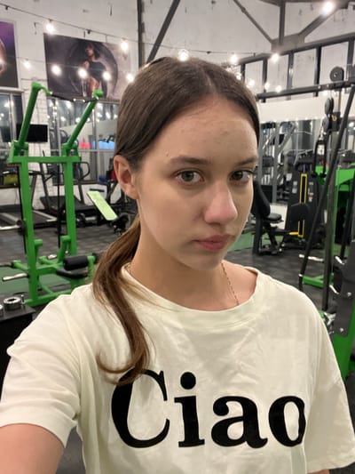 The scene is located indoors within a fitness center or gym in the city of Kozlovo, Russia. The primary subject is a young female, approximately late teens to early twenties, occupying the central and foreground portion of the image. She has dark brown hair, parted centrally and pulled back into a ponytail on her left side, with some loose strands framing her face. Her eyes are light brown or hazel and she looks directly into the camera with a neutral expression, her lips slightly parted in a subtle pout. Visible on her forehead are several small, raised skin blemishes. She wears a light-colored (off-white or very light yellow) short-sleeved t-shirt with the word "Ciao" printed in large black capital letters across the chest. A thin, silver-colored chain necklace is visible around her neck.
The background reveals a typical gym environment with various exercise equipment. To the left, a prominent green-framed resistance training machine is visible, likely a lat pulldown or similar apparatus. Further back on the left, a black treadmill and another workout bench are partially discernible. The wall on the far left features a white or light-colored brick texture, adorned with two large posters depicting muscular individuals; one is a male model positioned in front of a large tire, while the other poster is partially obscured. The ceiling is high, featuring exposed structural beams and decorative string lights with exposed bulbs, providing ambient illumination. To the right, dark-colored weight machines, including a cable machine, are visible, along with reflective mirrored walls that partially reflect other gym equipment. The flooring appears dark, consistent with gym surfacing. No other individuals are visible, and the woman is not actively engaged in exercise.