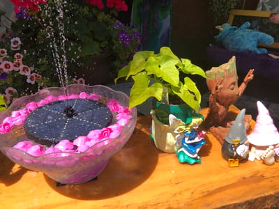 The image captures a close-up view of a wooden table with various decorative items, likely in an outdoor setting. In the foreground left, a clear glass bowl filled with water holds a black solar-powered fountain that is actively spraying water upwards. Pink artificial flowers are arranged around the rim of the bowl and floating on the water's surface. To the right of the fountain, a potted plant with bright yellow-green leaves is visible. Next to the plant, there is a decorative garden gnome holding a small yellow shovel, and a gnome with a pink hat. Further to the right, a decorative planter resembling a tree stump with a face is present, and behind it, a blue stuffed creature is visible. The background features an assortment of flowering plants with pink and purple blooms. The overall scene suggests a garden or a stall selling garden decorations. There are no people present in the image. The lighting indicates it is daytime with direct sunlight casting shadows on the wooden surface. The location is identified as the city of Severn, Canada.