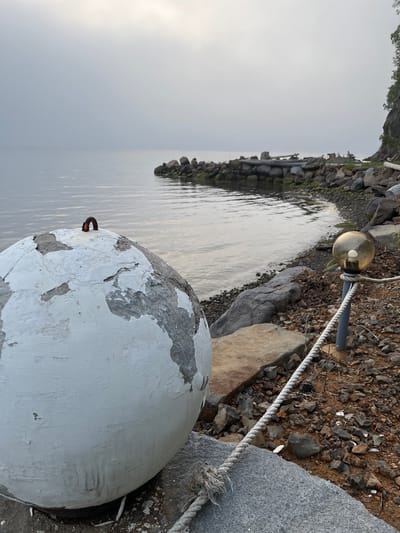 The image depicts a waterfront scene in Sovetskaya Gavan', Russia, under an overcast sky. In the foreground, a large, weathered, white spherical object, possibly a buoy, is positioned on a rocky surface. Its paint is peeling, revealing gray material underneath. A rusty metal ring protrudes from its top. To the right, a thin rope is secured to a metal post topped with a glass globe light fixture containing a visible bulb. The rope runs diagonally across the frame, towards the foreground. The ground is covered in small rocks and debris. In the middle ground, the calm water of the bay stretches towards a rocky shoreline. This shoreline is punctuated by large stones and patches of green moss or algae. The background is dominated by a soft, hazy sky and a distant, wooded hillside on the right.