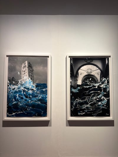 Two framed artworks are displayed on a textured, off-white wall. The artwork on top features a black and white image of an ornate circular architectural interior, possibly a rotunda with a chandelier, partially obscured by dark, abstract paint splatters and streaks of white and light blue. The painting appears to be abstract expressionist in style, with thick, impasto brushstrokes.
The artwork below is also a framed photograph with added paint. It depicts a building, likely in an urban setting, with numerous windows and architectural details, appearing to be submerged or impacted by a dramatic, turbulent wave rendered in deep blues, white, and black paint. The wave is thick with texture, suggesting movement and force. The background sky is overcast and gray, with some scattered white specks that could be rain or spray.
The overall presentation suggests an art exhibition. The lighting creates soft shadows on the wall, indicating the artworks are illuminated. There is no visible text within the artworks or on their frames. The style of the artworks suggests a contemporary art context.