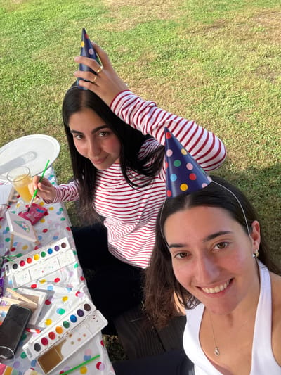 Two young adult females are seated outdoors at a table engaged in an art activity, identified as being in Accord, United States. The background consists of manicured green grass under natural light. The female on the left wears a red and white striped long-sleeved shirt and has dark hair; she holds a dark blue polka-dotted party hat with her right hand, displaying a gold ring on her ring finger, and holds a green-handled paintbrush in her left hand, which is immersed in an orange liquid in a clear glass. She looks towards the camera with a slight smile. The female on the right wears a white halter-neck top and has dark hair; she wears a similar dark blue polka-dotted party hat secured by a white headband, a small gold earring in her left ear, and a silver pendant necklace. She smiles broadly, looking directly at the camera. The table is covered with a white polka-dotted tablecloth, holding multiple white watercolor paint palettes with various colored paints, several small blank canvases, additional paintbrushes, and a black smartphone.