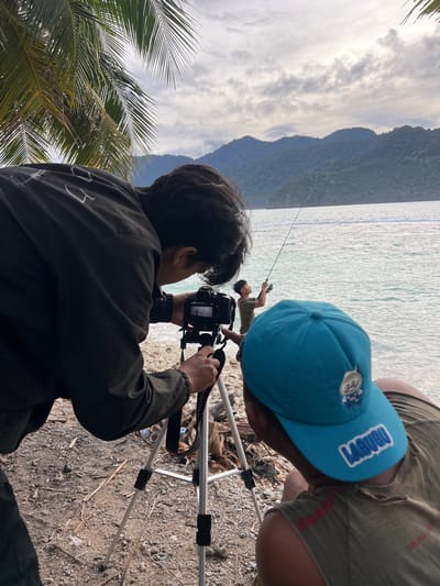 In Babahie, Indonesia, two individuals are positioned on a rocky shore by the sea. The individual in the foreground, wearing a turquoise cap with a cartoon character and the word "LABUBU" emblazoned on it, is facing away from the camera, looking towards the water. To their left, another person, dressed in a dark jacket, is operating a camera mounted on a tripod. This person is focused on capturing an image of a third individual further out in the water. This third person, wearing a green t-shirt, is actively fishing with a rod. The backdrop consists of a calm sea and a lush, tree-covered mountain range under an overcast sky. Palm fronds from a nearby tree are visible in the upper left corner of the frame.