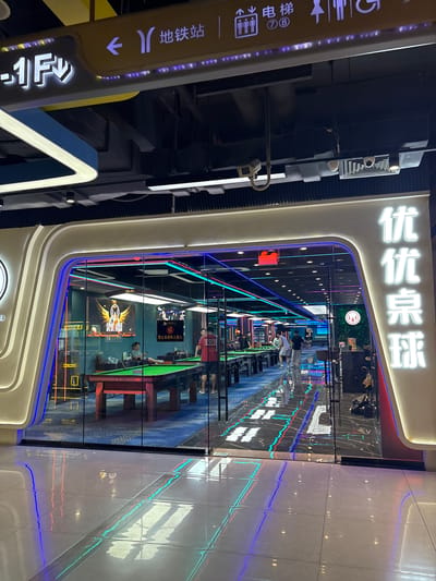 The image shows the exterior of a billiards hall in Haizhu District, China. The entrance is a large, brightly lit doorway with a curved white frame and neon blue and red accents. Through the glass doors, multiple pool tables are visible, illuminated by colorful neon lights. People can be seen playing billiards inside. The floor in front of the entrance is tiled and reflects the neon lights from the establishment. Overhead signage indicates directions to "Subway Station" and "Elevator." The overall impression is a modern and vibrant entertainment venue.