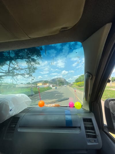 The image is a photograph taken from inside a vehicle looking out through the windshield. The view shows a road with trees and buildings on either side. In the distance, a large rock formation is visible. The sky is blue with some clouds.

There are two decorative balls hanging from the interior of the car. One is orange and the other is pink. A roll of plastic wrap is also visible on the dashboard.

The location is identified as Ikere Ekiti, Nigeria. The scene depicts a typical day in a Nigerian town with a road, vegetation, and residential buildings. The rock formation in the background is a prominent geographical feature. The presence of the balls and plastic wrap suggests personal touches within the vehicle. The overall impression is one of everyday life in this Nigerian city.