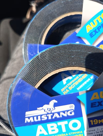 The image displays a close-up, top-down perspective of several rolls of black double-sided foam adhesive tape, situated in what is identified as Maiaky, Ukraine. Each roll features a blue and light blue label. Prominently displayed on the labels are a white stylized horse logo and the brand name "MUSTANG" in white capital letters, with "АВТО" (AUTO) visible below. Additional Ukrainian text on the labels reads "ДВОСТОРОННЯ КЛЕЙКА СТРІЧКА НА ВСПІНЕНІЙ ОСНОВІ" (Double-sided adhesive tape on a foamed base) and partial dimensions "19MM X 5M". The tape exhibits a black foam core with thin, light-colored adhesive lines visible on the edges of the rolls, wound around a light brown cardboard core. The immediate background on the left consists of blurred grey fabric, consistent with upholstery or clothing, while the upper background is indistinct and dark. Direct, natural light illuminates the objects from the upper right, casting soft shadows. No individuals are present in the frame.