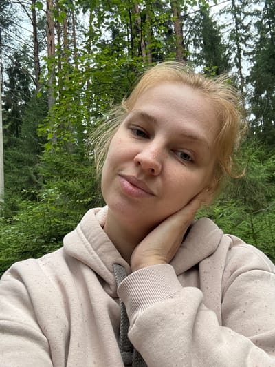 A young Caucasian woman with blonde hair and fair skin with freckles, wearing a light pink hooded sweatshirt, is pictured in a selfie outdoors. She has her right hand supporting her chin and cheek. Her eyes are partially closed, and her lips are slightly parted, giving her a relaxed or pensive expression. The background consists of a dense forest with tall trees and lush green foliage, indicative of a natural, wooded environment. The lighting suggests it is daytime, possibly overcast or shaded by the trees. The image appears to be taken at an outdoor location, possibly a park or a trail within or near the city of Zaymishche, Russia, given the dense forest setting.