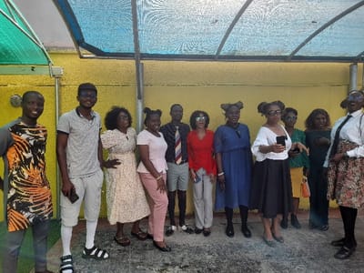 The image depicts a group of eleven individuals, consisting of three males and eight females, posing outdoors in the city of Ajah, Nigeria. They stand against a solid yellow wall, beneath a translucent blue and green mesh or corrugated plastic canopy supported by metal beams. The ground is a dark, possibly concrete or asphalt, paved surface.
From left to right:
1. A male in a grey t-shirt with orange and black patterns and grey trousers.
2. A male wearing a dark cap, sunglasses, a grey polo shirt, light shorts, white socks, and black and white slide sandals, holding a dark smartphone.
3. A female with curly hair and glasses, wearing a light-colored patterned dress, with her left hand on her hip.
4. A female with dark hair styled in two buns, wearing a light pink top and light pink trousers.
5. A male with a light blue shirt, a striped tie, and light grey shorts.
6. A female with dark curly hair and sunglasses, wearing a red long-sleeved top and light grey trousers, holding a small white object.
7. A female with dark hair in two buns, wearing a dark blue long-sleeved dress.
8. A female with dark hair in two buns and sunglasses, wearing a white off-the-shoulder blouse and a black pleated skirt, holding a dark smartphone and gesturing with her left hand.
9. A female with dark curly hair and sunglasses, wearing a dark green long-sleeved top and a yellow/orange patterned skirt, holding a yellow bag.
10. A female with dark hair and glasses, wearing a dark green long-sleeved top and dark trousers.
11. A female with dark hair and glasses, wearing a white collared shirt, a dark tie, and a patterned knee-length skirt.
Most individuals are smiling and looking towards the camera. Sunlight filters through the canopy, illuminating the scene.