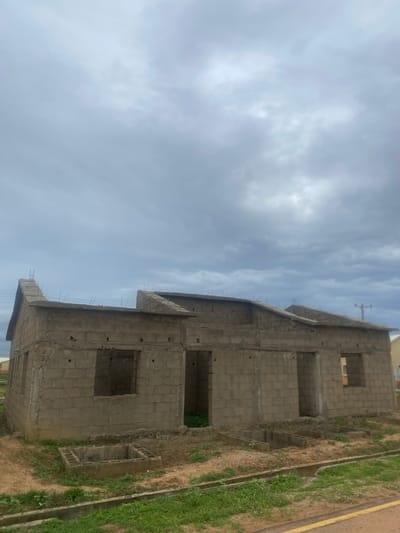 The image shows the partially constructed exterior of a building made of concrete blocks. The structure has multiple stories with unfinished windows and openings. Some rebar protrudes from the concrete edges, indicating ongoing construction.
The scene is outdoors, with a cloudy sky overhead. The ground around the building is covered in dry earth and patches of green grass, with a narrow drainage ditch visible. A yellow line marking is present on the right side of the image, possibly indicating a road or boundary.
No people or specific activities are visible, suggesting the construction is paused or the image was taken from a distance. The lighting suggests it could be daytime, with diffused light due to the cloud cover. There is no visible text in the image. The location context is Bulamare, Nigeria.