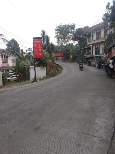 The image depicts a paved road winding through a semi-urban area, likely in Megamendung, Indonesia. On the left, a white building with red trim and prominent windows is visible, adjacent to lush green foliage and a RedDoorZ sign displaying amenities like free WiFi, mineral water, television, and air conditioning. Further down the road, a red building with multiple windows stands out against the greenery.
Several motorcycles are present. Two people are riding on one motorcycle traveling away from the viewer. Another motorcycle is parked on the right side of the road, near a small roadside stall. Several other motorcycles are parked further down the road. Two individuals, one in a green shirt and another in a white and brown outfit, appear to be standing near the stall. The road surface is concrete with visible expansion joints and some staining. Overhead power lines are strung across the sky. The overall environment suggests a residential or commercial area nestled amongst tropical vegetation.