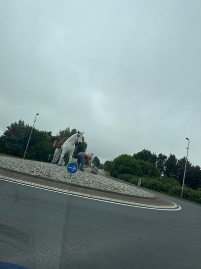 The image depicts a roundabout in the city of Le Tilleul, France. A large, white statue of a horse with saddlebags is the central feature, positioned on a gravel-covered island. A human figure, dressed in a red shirt and blue jeans, is bent over near the horse's hindquarters, appearing to be engaged in an activity related to the statue or the surrounding area. A blue circular sign with a white arrow pointing to the right is visible on the roundabout. The background consists of green trees and a cloudy, overcast sky. The foreground shows a section of asphalt road, indicating the perspective is from a vehicle.
